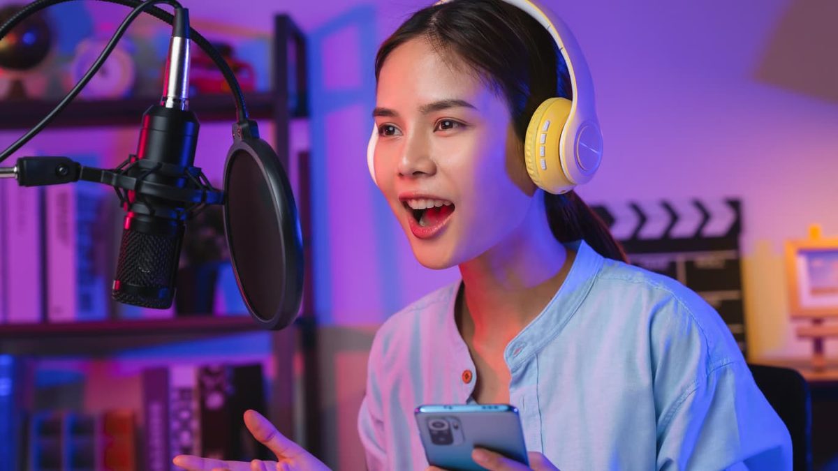 A host in headphones speaking into a microphone in a studio lit pink and blue