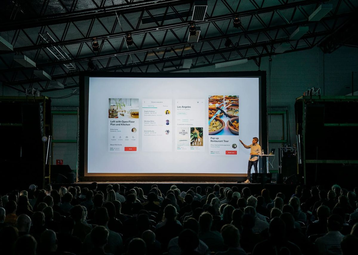 A speaker presenting slides to a large seated audience in a dark hall