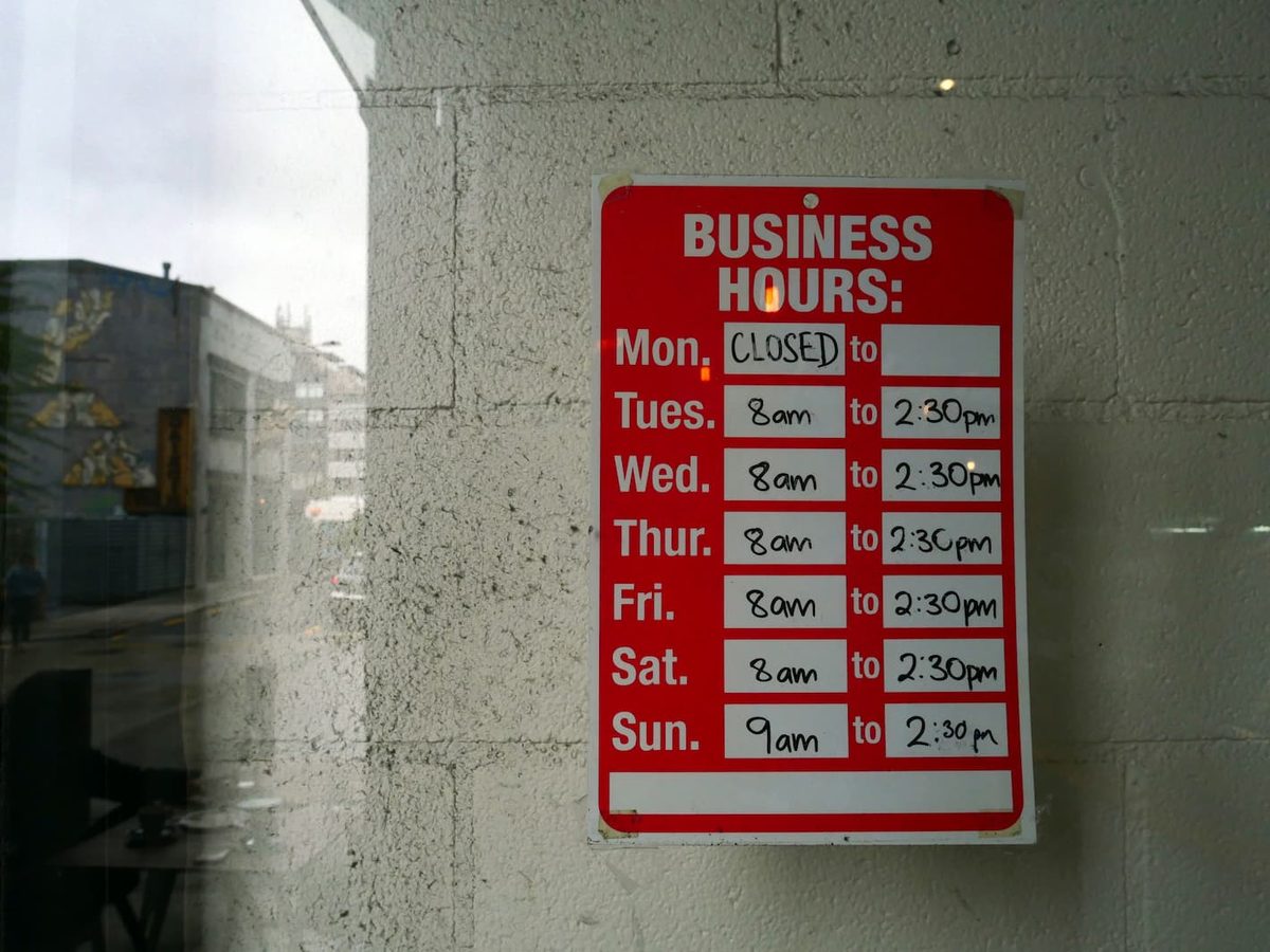 A red business hours sign on a concrete wall, listing opening times for each day