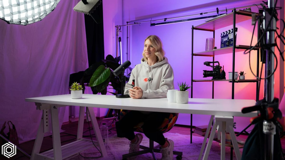 A host at a desk in a studio lit pink and purple, surrounded by cameras and lights